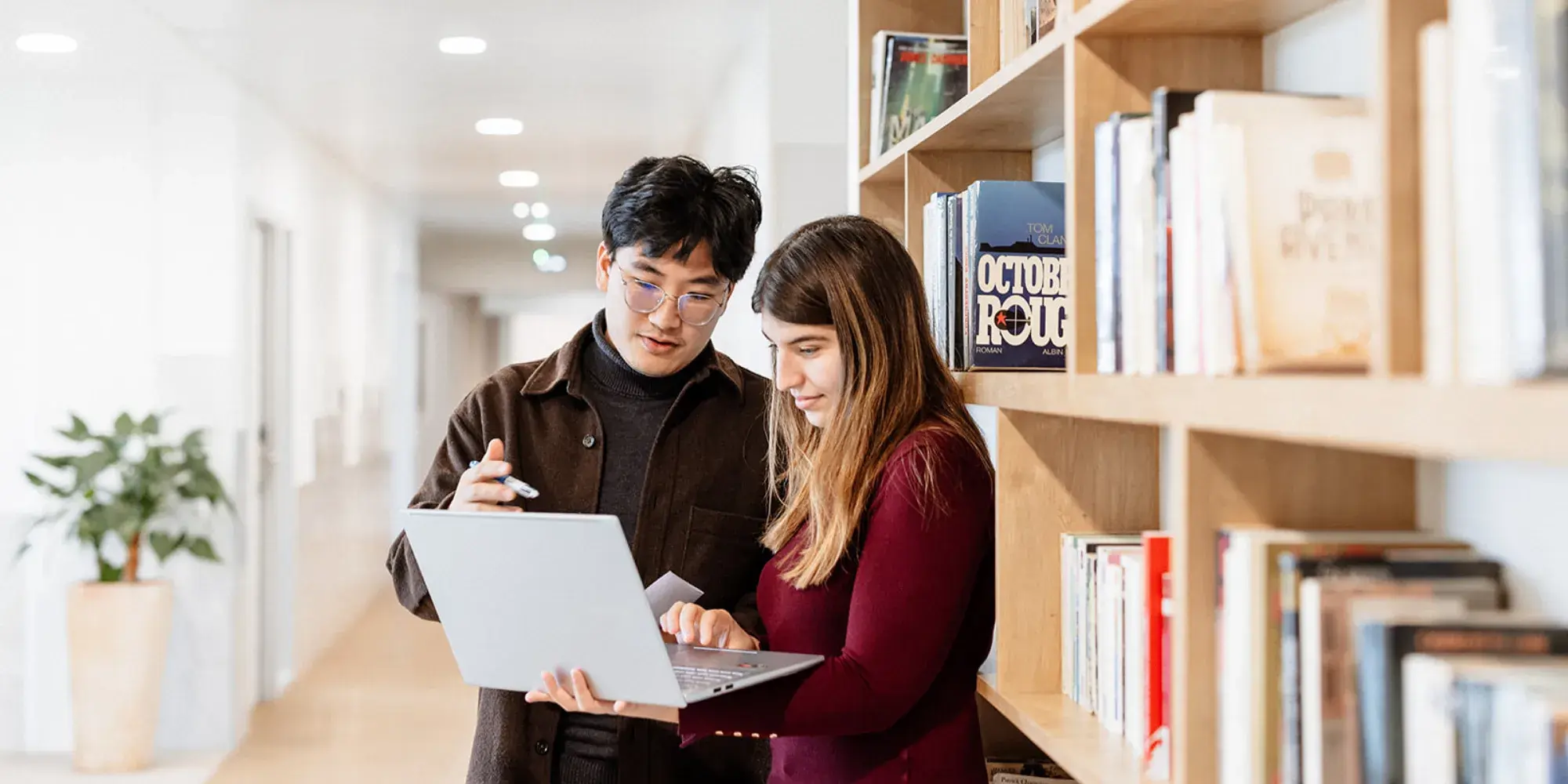 Deux étudiants de SKEMA debout devant une bibliothèque, regardant ensemble l’écran d’un ordinateur portable.