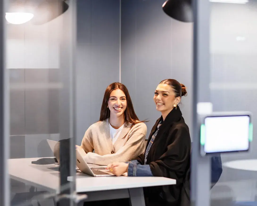 Deux jeunes filles souriantes regardant l'objectif, assises devant un ordinateur.
